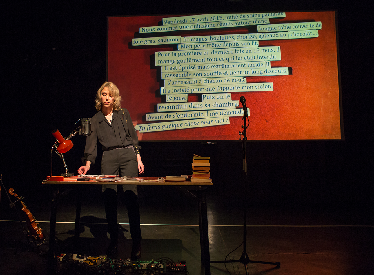 Blond woman stands at a desk with lamp and books in front of a projection of French writing.