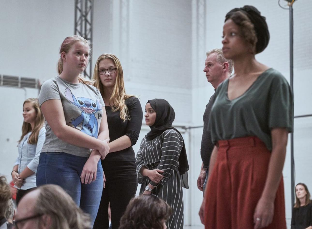 A group of people stand and sit in a white rehearsal room.