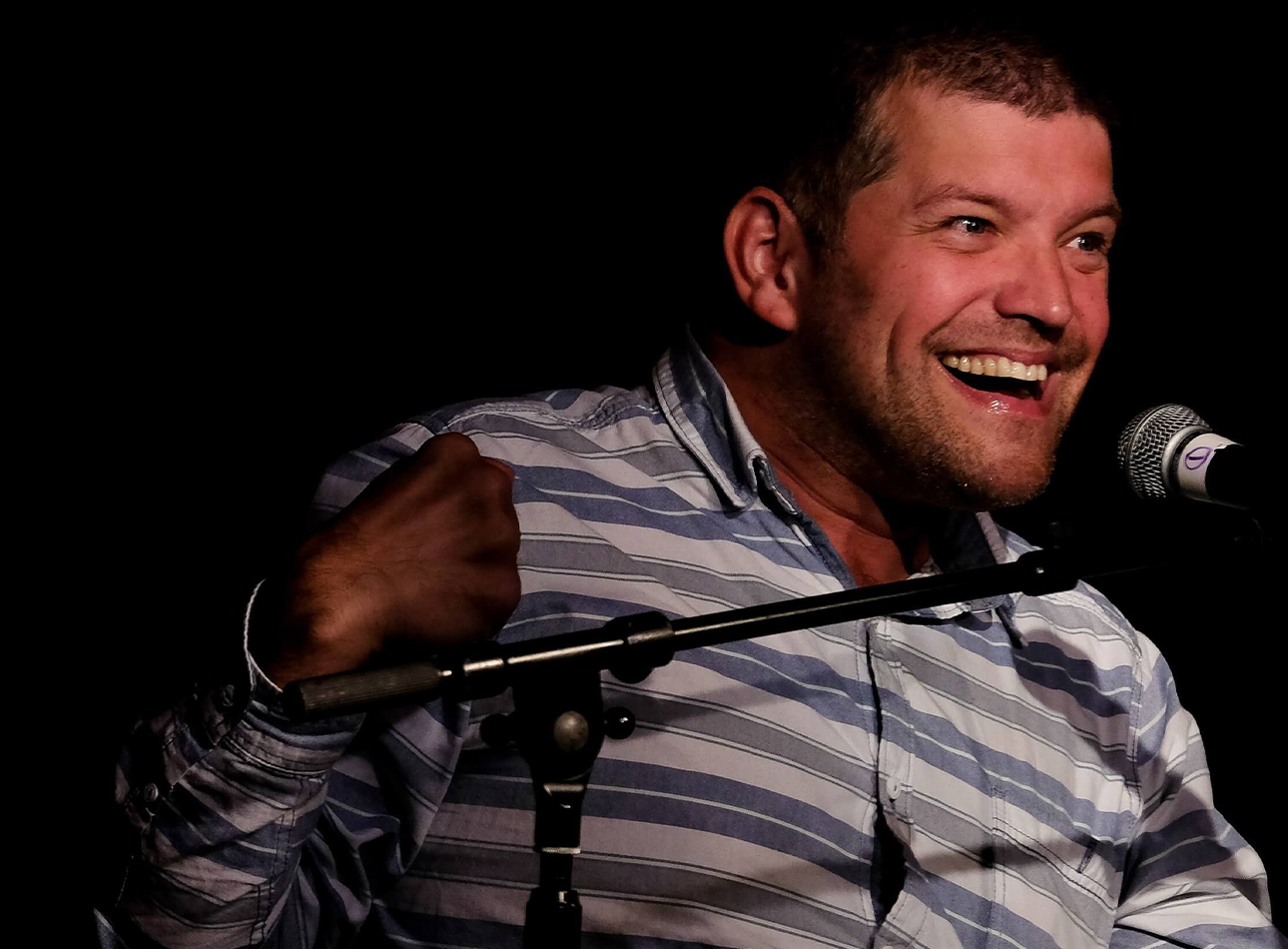 A man smiles into a mic, wearing a blue and white striped shirt.