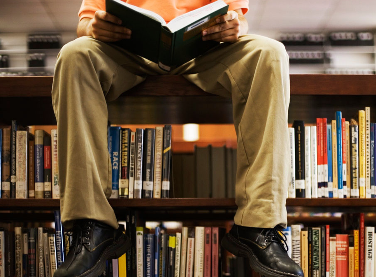 A bookcase with a person sat with an open book on top, legs dangling down. Face is not visible.