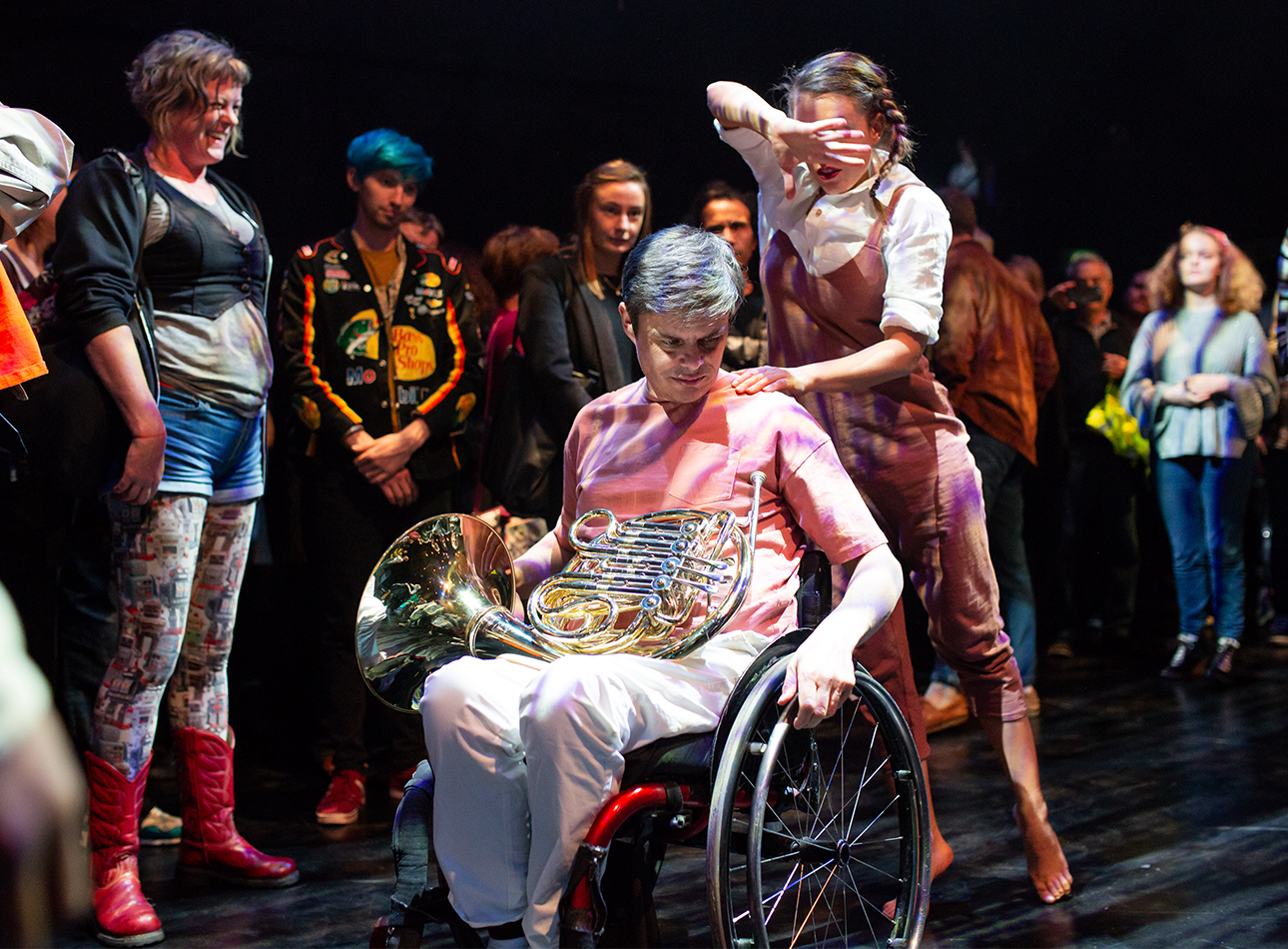 A man in a wheelchair holds a French Horn in a circle of diverse people in casual dress.