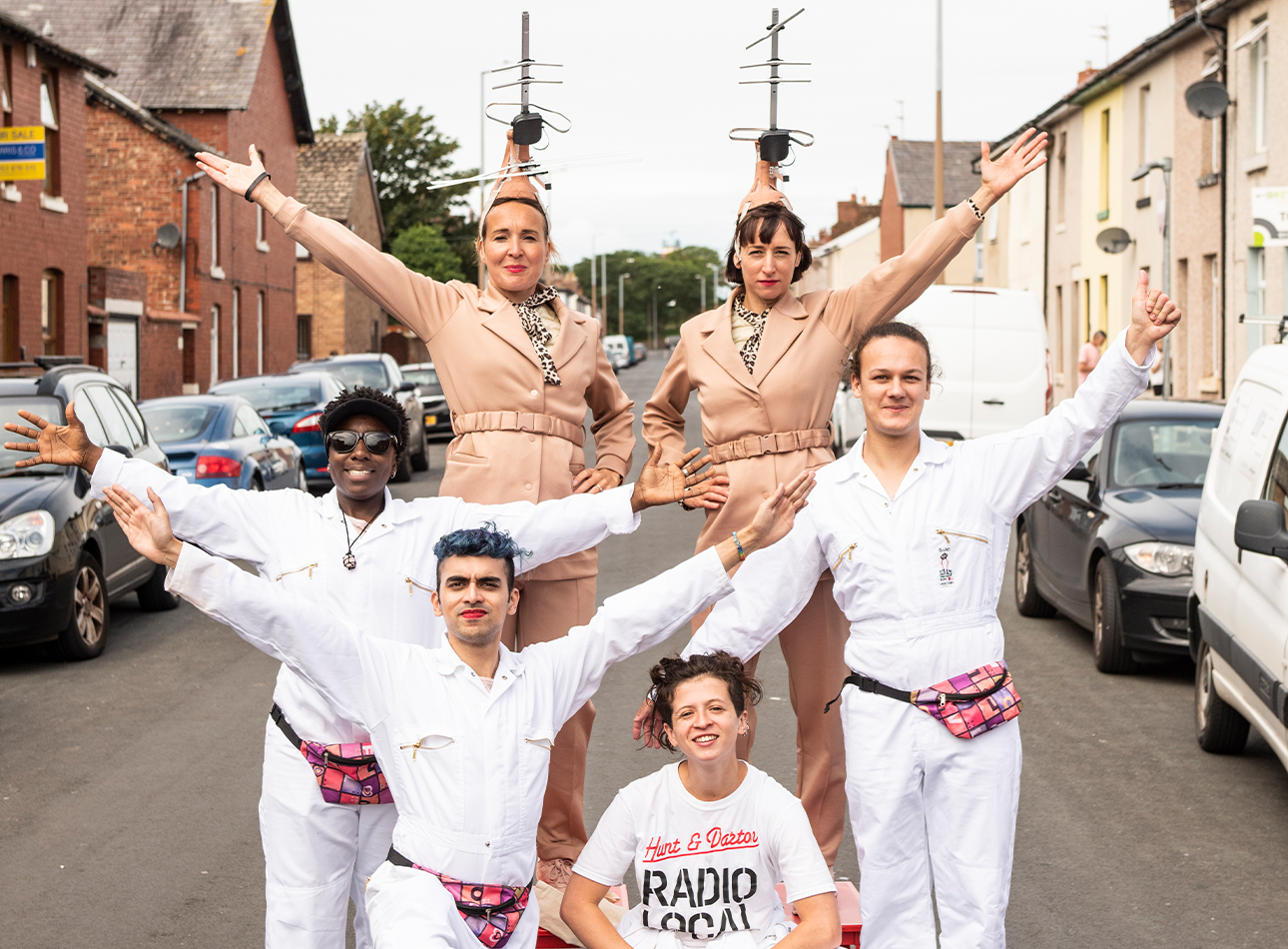 4 people in white jumpsuits and 2 people in beige jumpsuits stand with their arms raised in the middle of an estate.