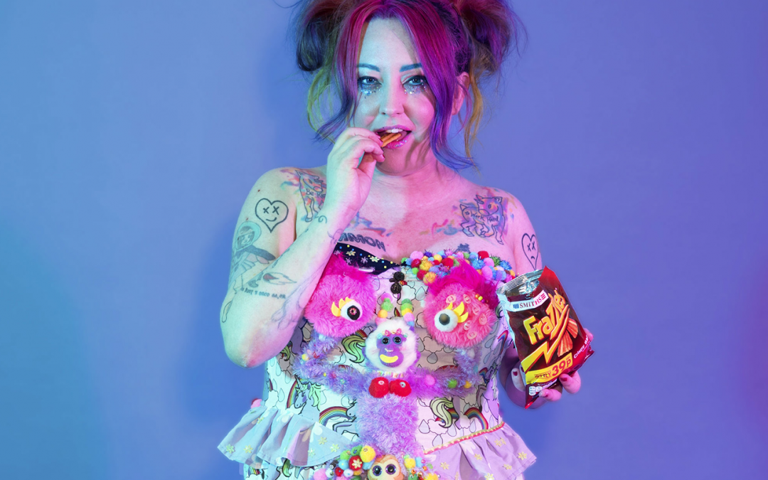 A woman with tattoos and colourful hair eats a packet of Frazzles in an expressive, multicoloured dress adorned with a purple monkey.