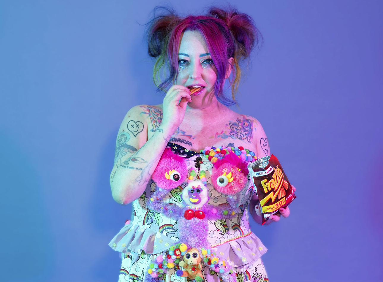 A woman with tattoos and colourful hair eats a packet of Frazzles in an expressive, multicoloured dress adorned with a purple monkey.