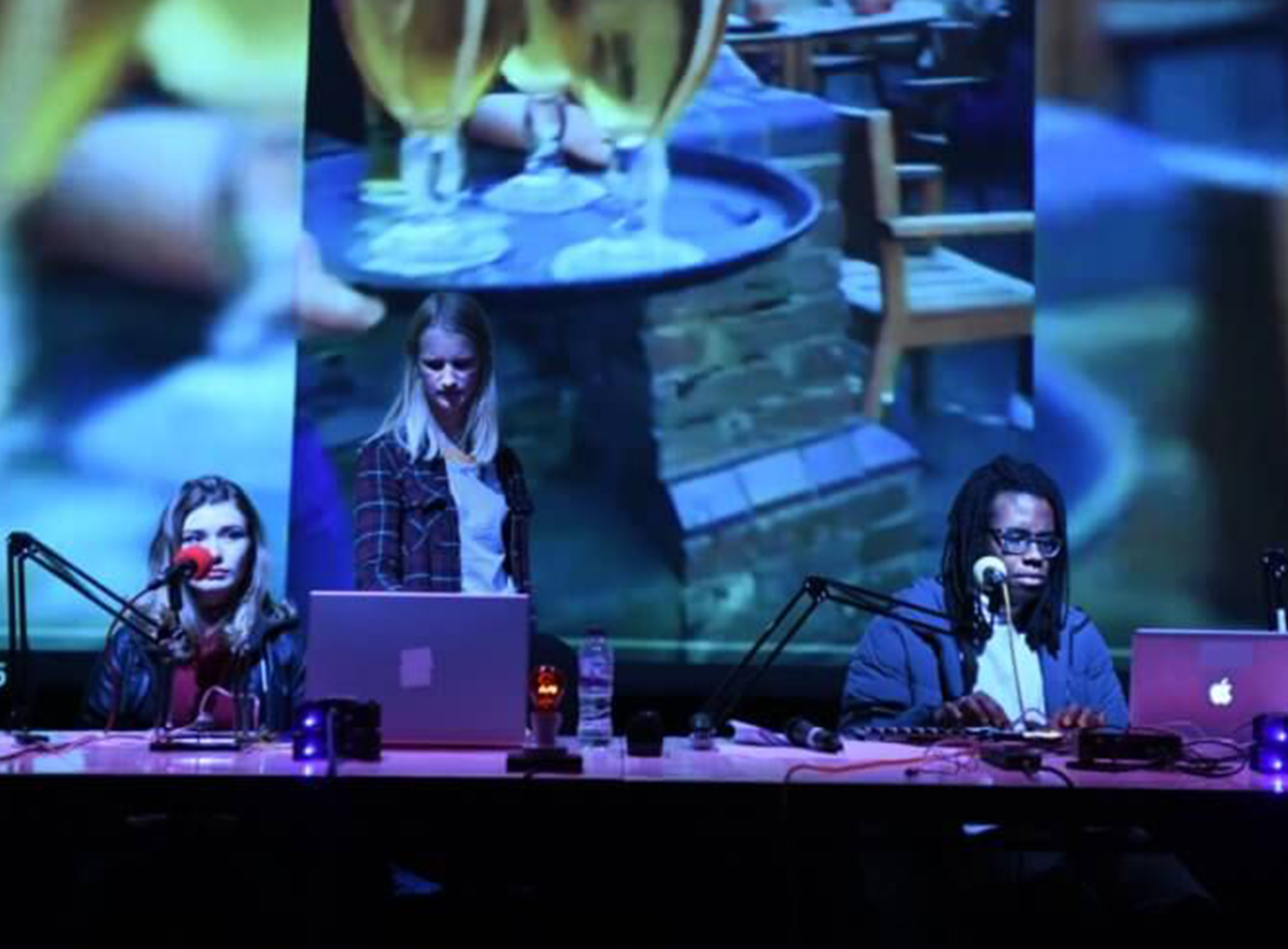 A purple and blue washed image of 2 people sat at a desk with computers and microphones in front of a projection of a pub.