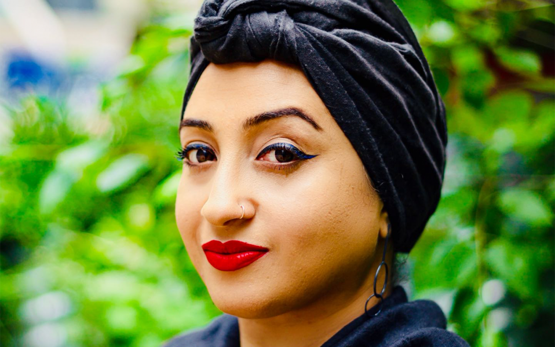 A woman with black winged eyeliner and red lipstick wearing a black turban and black shawl.