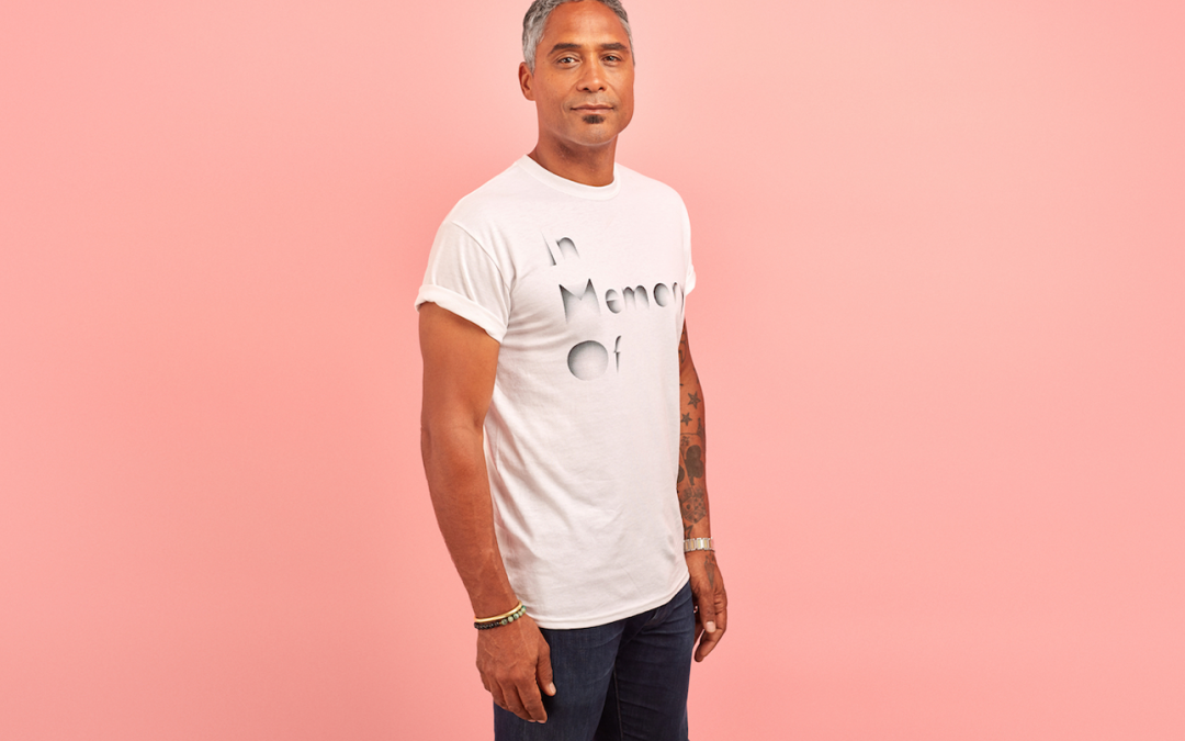 A man with grey hair wears a white t shirt that reads 'In Memory Of', against a pink background.