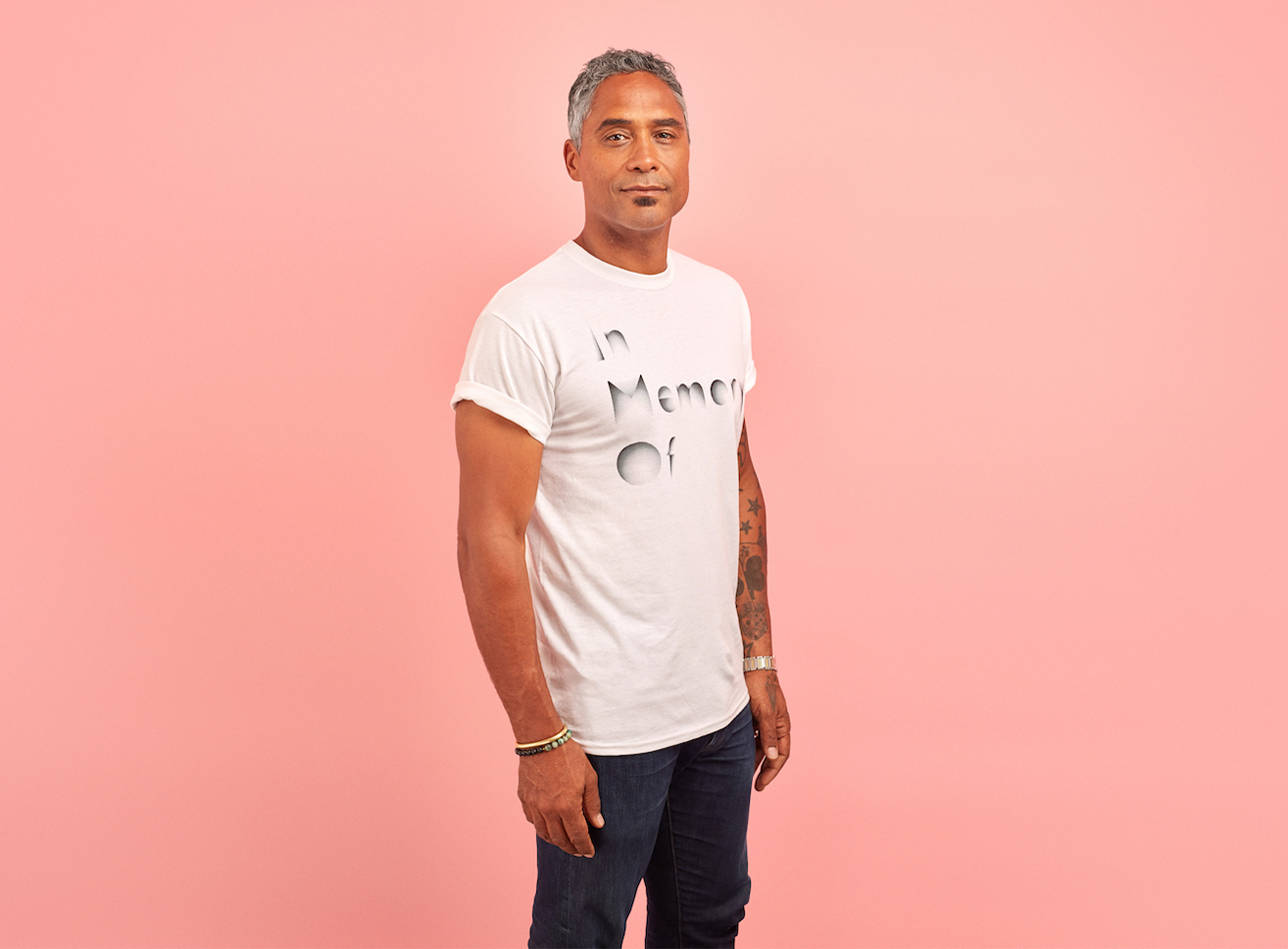 A man with grey hair wears a white t shirt that reads 'In Memory Of', against a pink background.