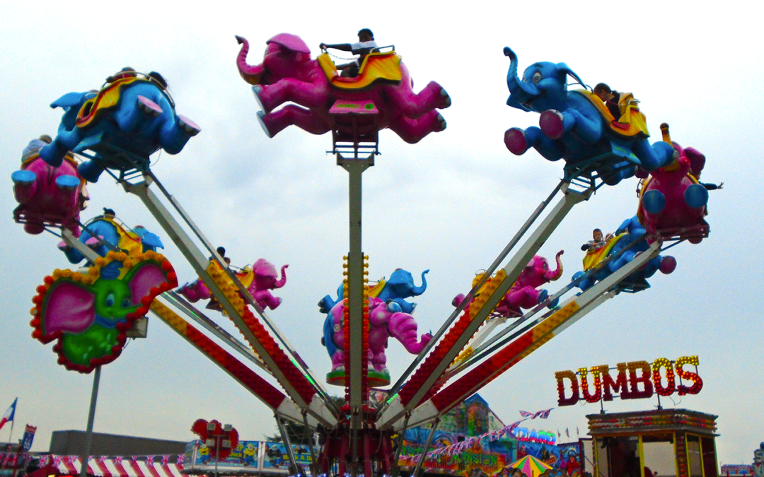 An elephant themed fair ground ride up in the air, with a fair in the background
