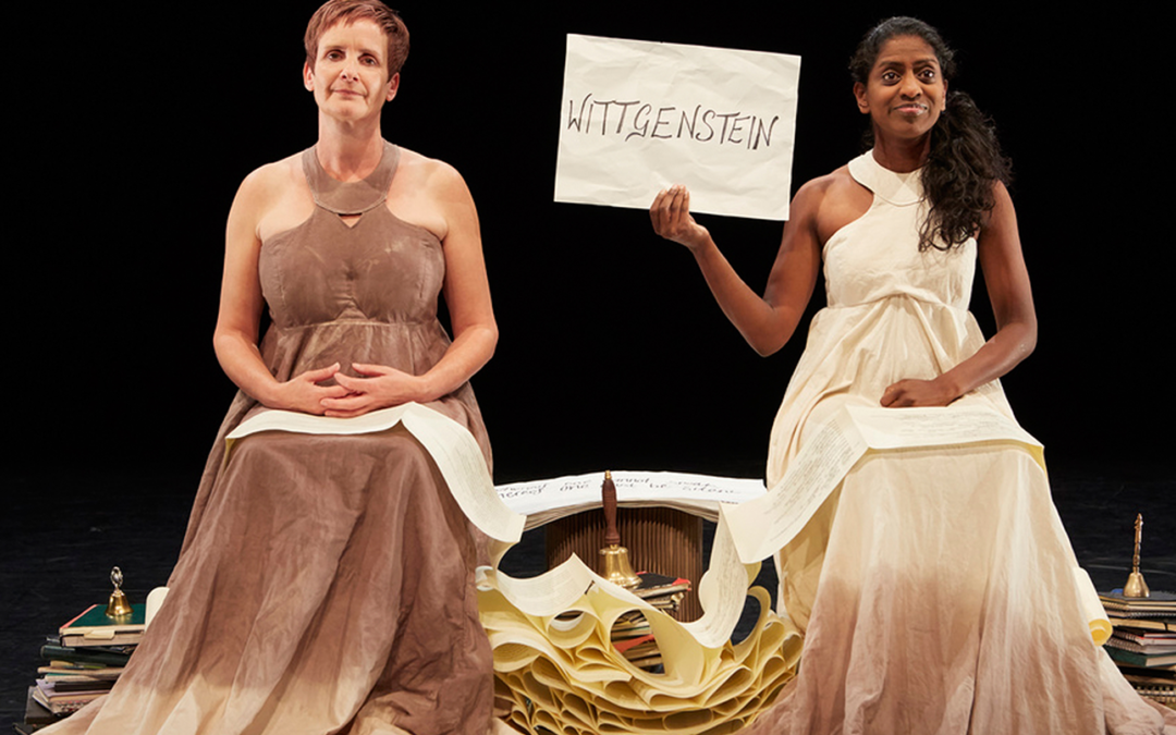Two female performers sat next to another in neutral tone dresses, the performer on the right holds a sign that reads 'Wittgenstein'