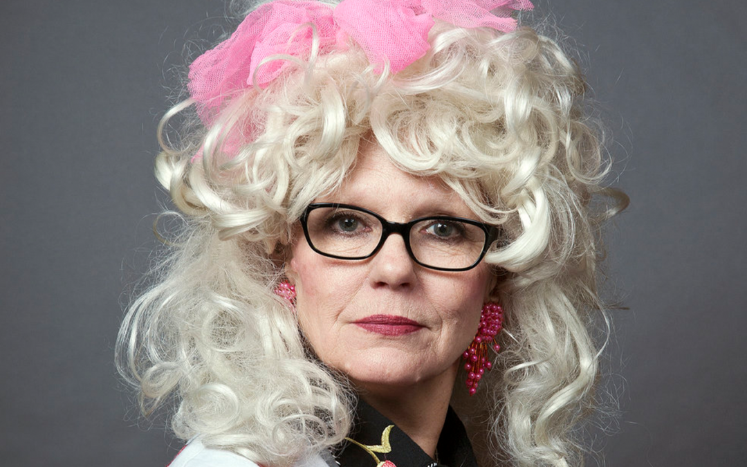 Tammy WhyNot looks directly at the camera wearing glasses, a big blonde curly wig with a pink scrunchie.