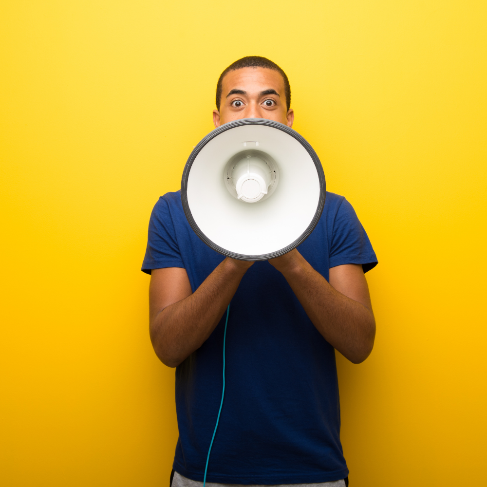 A young man in a dark blue shirt is holding a white megaphone. He is standing in front of a bright yellow background