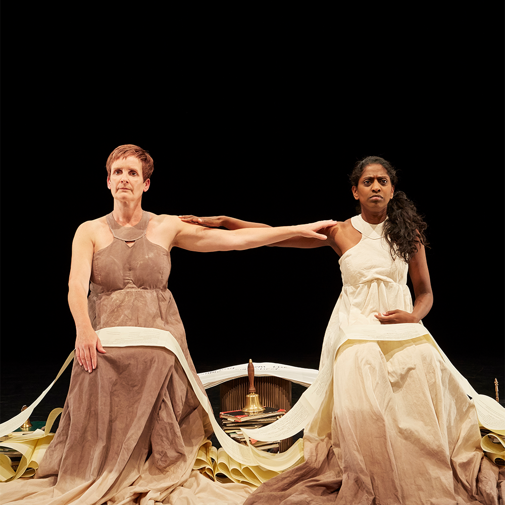 Two female performers sat next to another in neutral tone dresses holding eachothers shoulder as they look out