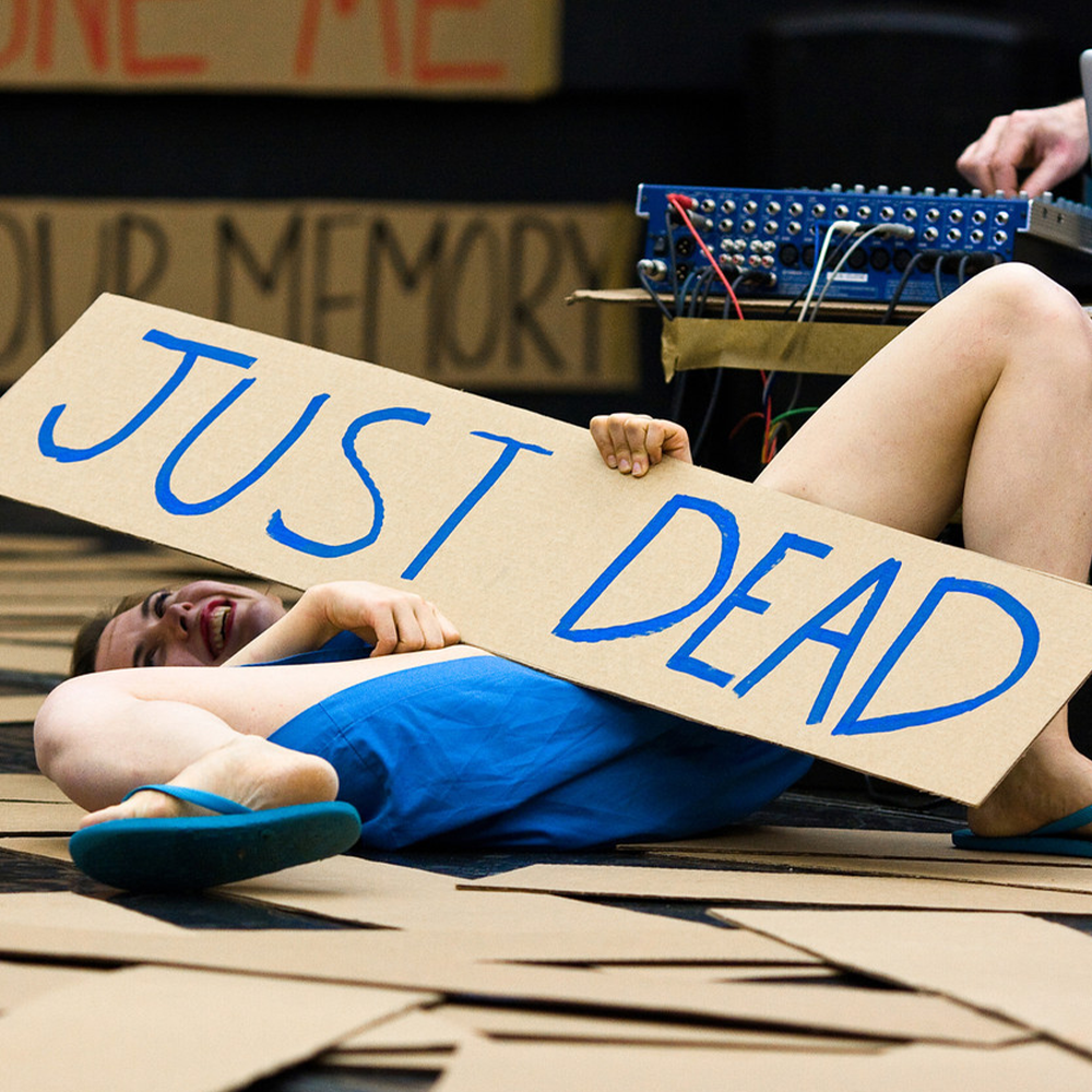 Woman in blue dress and flip flops surrounded by placards holding a placard that reads 'Just Dead'