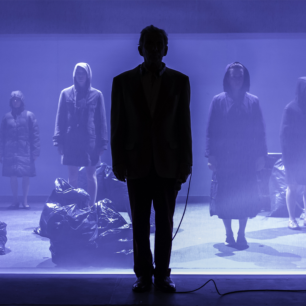 Stage washed in purple, people stood in background holding luggage with silhouette of man in centre