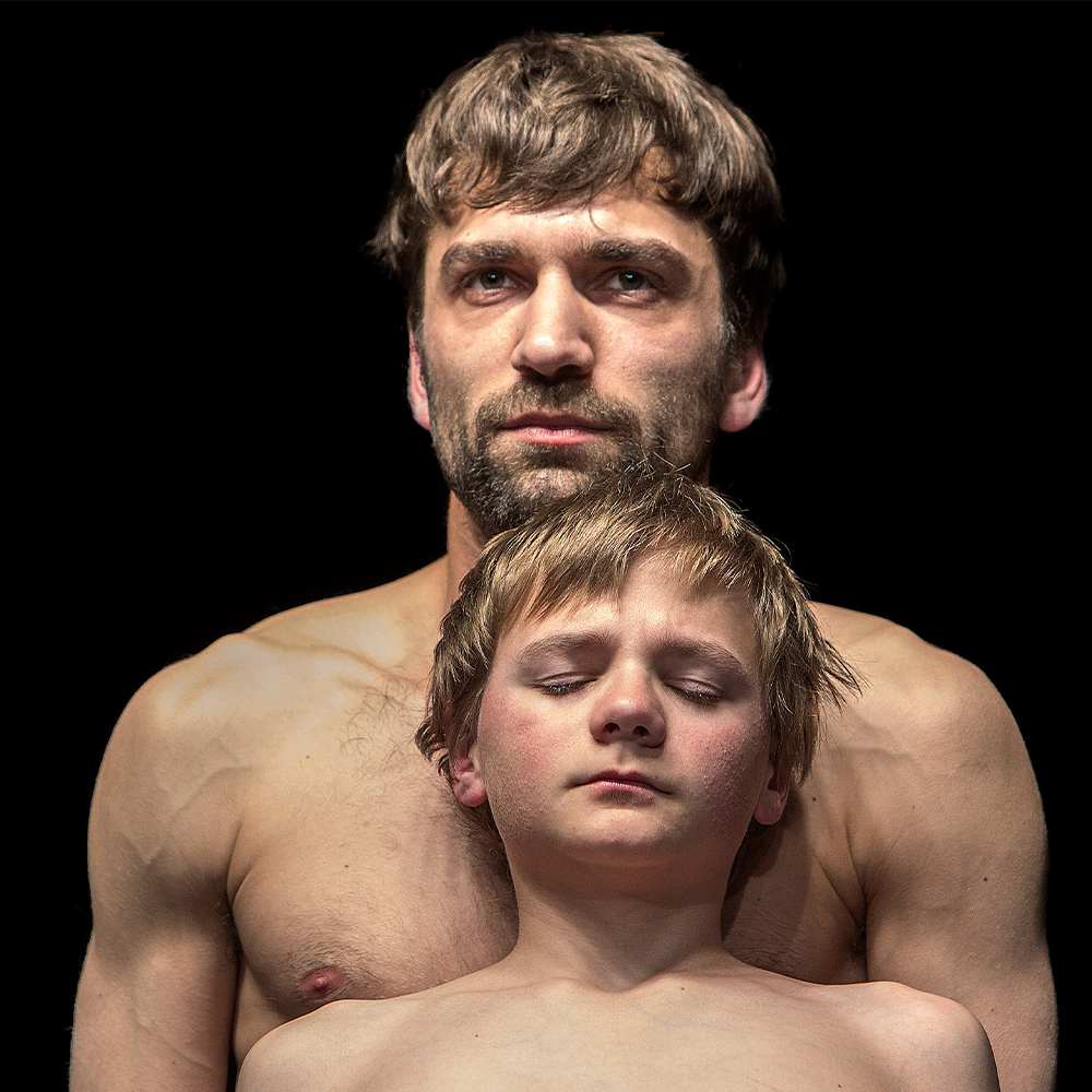 Both performers stood topless against a black background. The young performer in front has his eyes closed.