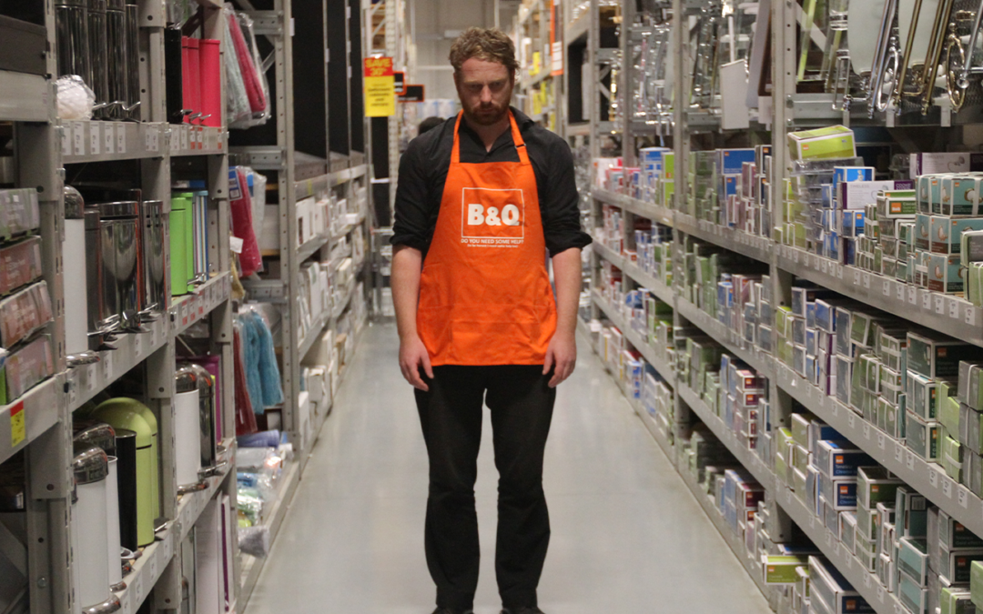 A man stood in a warehouse wearing an orange apron that reads 'B & Q'