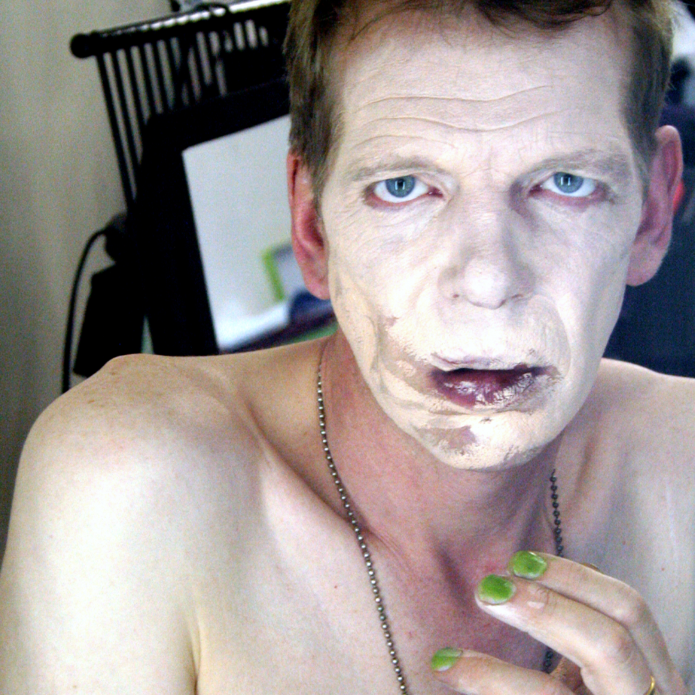 Topless man looking down the camera with smeared light makeup covering his face