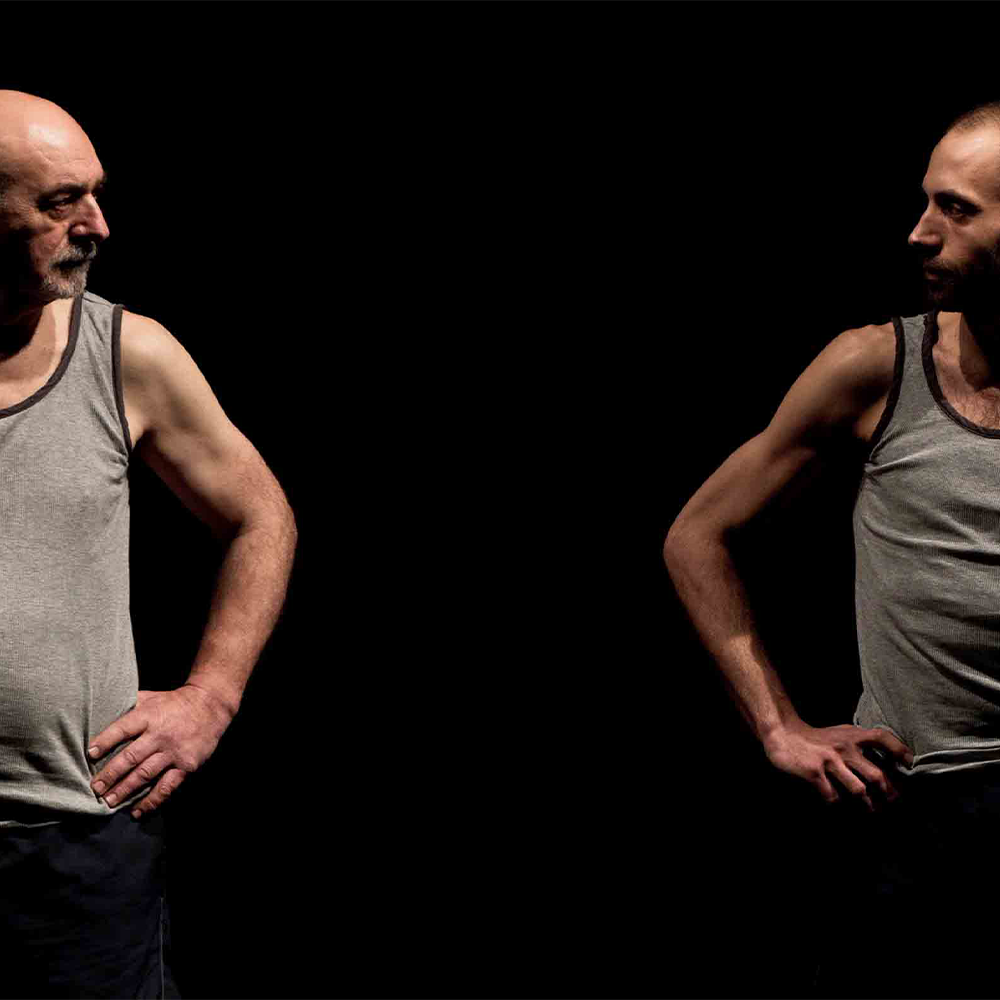 Two men in grey vests look at one another with hands on their hips