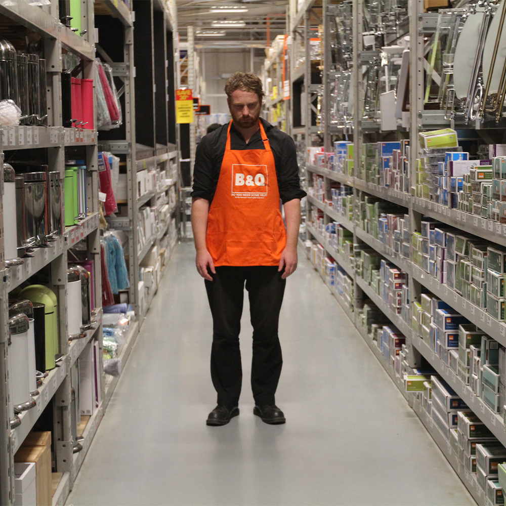 A man stood in a warehouse wearing an orange apron that reads 'B & Q'