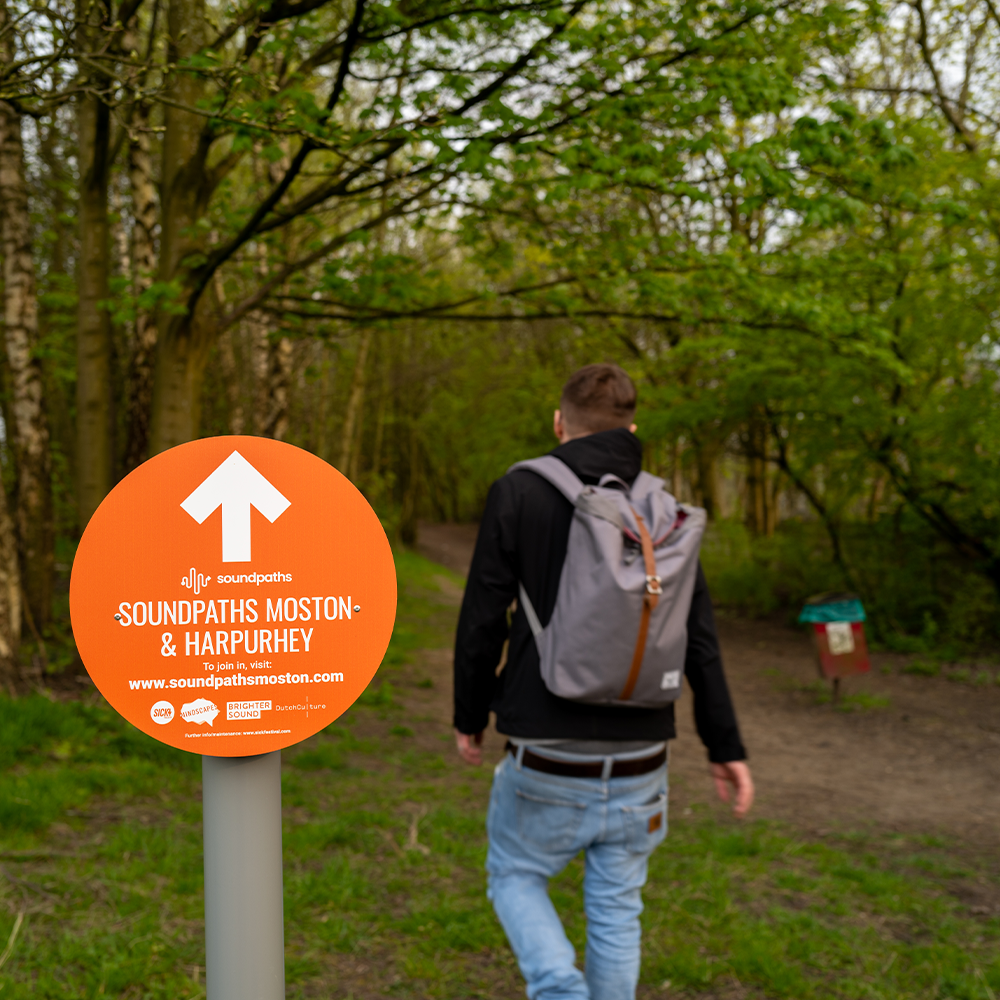 An orange sign reads 'Soundpaths' as a man is seen walking the route in nature.