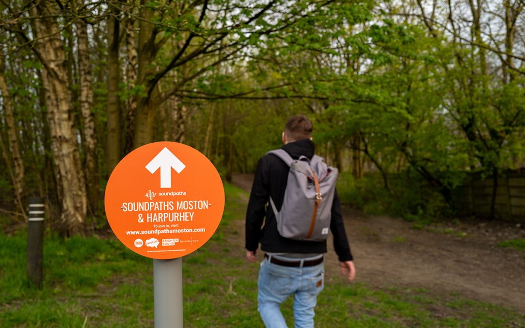 An orange sign reads 'Soundpaths' as a man is seen walking the route in nature.