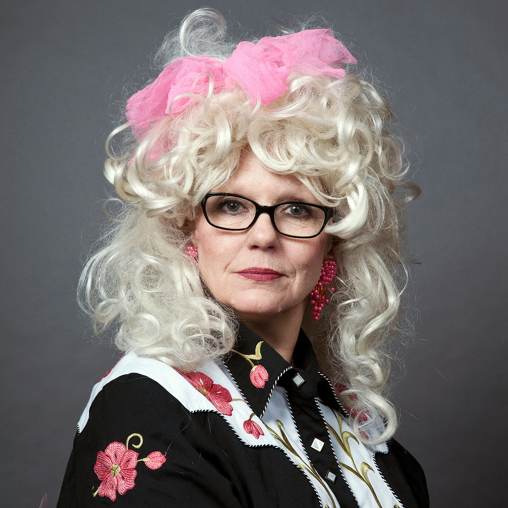 Tammy WhyNot looks directly at the camera wearing glasses, a big blonde curly wig with a pink scrunchie.