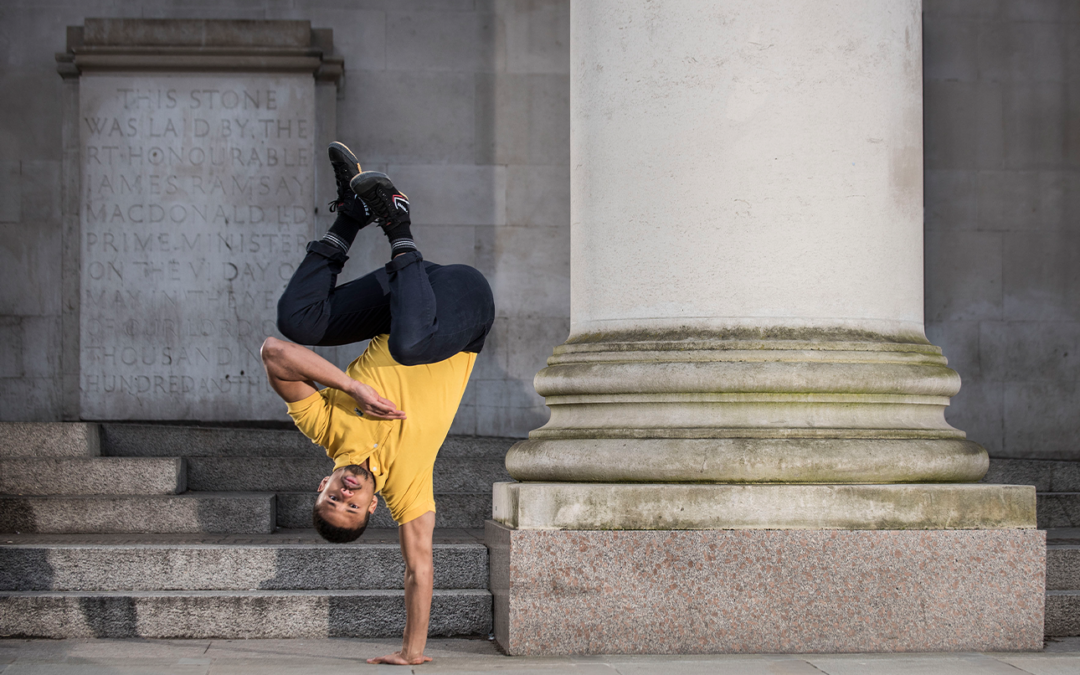 A man in yellow t shirt and blue jeans does a one handed handstand