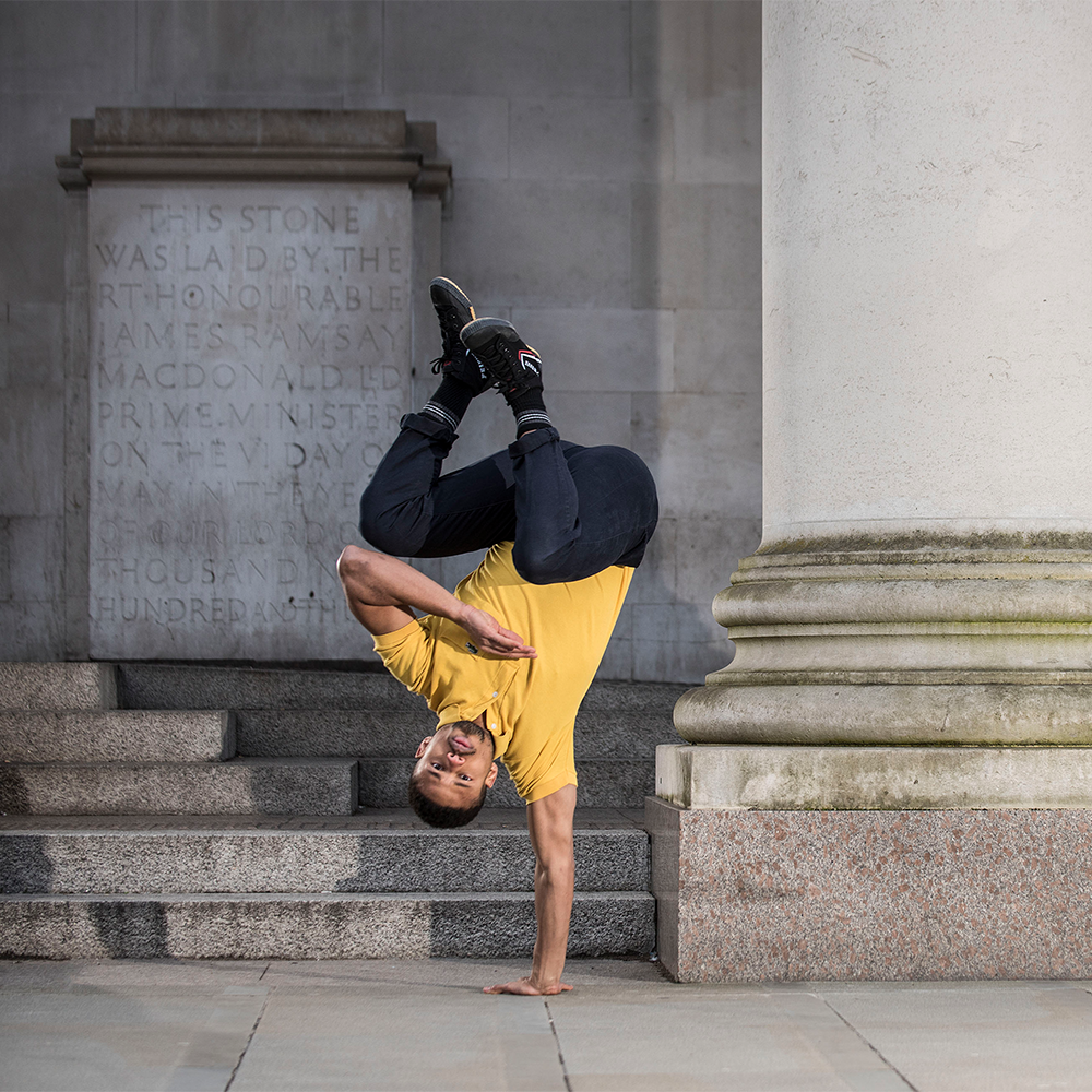 A man in yellow t shirt and blue jeans does a one handed handstand